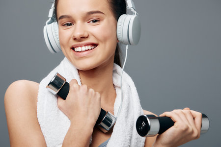 Woman fitness portrait of a smiling young woman holding dumbbells with a towel draped over her shoulders and headphones on, sport workout concept in a grey studio background.の写真素材