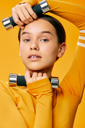 Young woman fitness portrait holding chrome dumbbells near her face, wearing a yellow long-sleeve top against an orange studio background, close-up with confident expression and glowing healthy skin.の写真素材