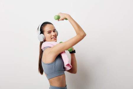 Young woman fitness portrait lifting green dumbbells while wearing headphones, gray sports bra, pink towel over shoulder, smartwatch visible, smiling against white background.の写真素材