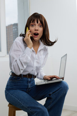 Young woman sits with laptop, talking on a phone, eyes wide in surprise. Bright natural light and...