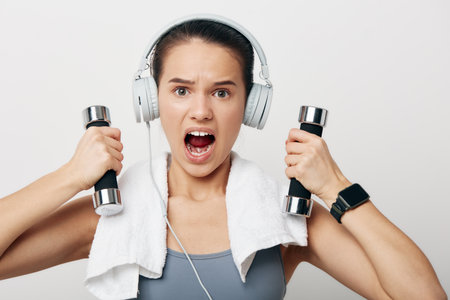 Young woman fitness portrait lifting chrome dumbbells with headphones and towel, intense expression,...