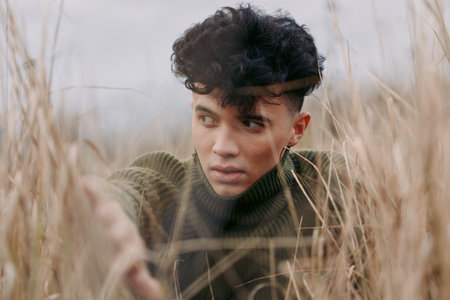 A candid portrait of a young man peering through tall dry grass, captured with natural light and...
