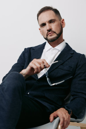 A composed business man in a textured blazer sits against a plain light background, projecting confidence and focus while adjusting his glasses.の写真素材
