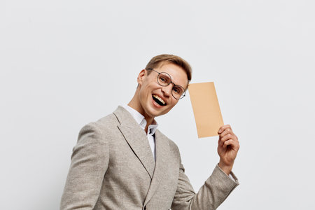 Cheerful young man in glasses smiling broadly while holding blank cardboard sign, dressed in stylish beige checkered blazer, white shirt, isolated on plain gray background, studio portrait. People lifestyle conceptの写真素材