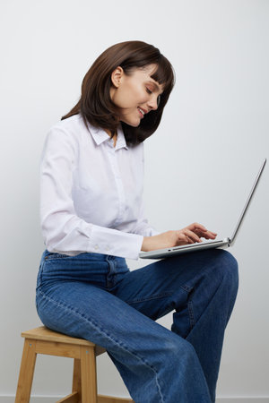 A focused woman sits on a wooden stool, typing on a slim laptop. She wears a white shirt and blue jeans, projecting casual professionalism in a bright, uncluttered studio setting.の写真素材