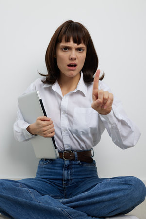 Young woman sits crosslegged against a plain wall, holding a laptop and raising a finger with a firm, commanding expression, signaling warning, instruction, or decisive talk.の写真素材