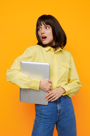 A surprised young woman in a yellow shirt and jeans clutches a laptop against a bright orange background, capturing a moment of curiosity, surprise, and casual tech workflow.の写真素材