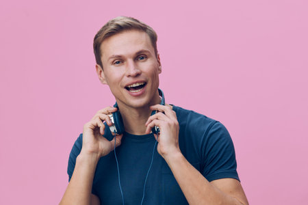 Upbeat young caucasian man wearing casual blue t-shirt and headphones, smiling happily while enjoying music, standing against pastel pink background. People leisure lifestyle concept, positive emotionの写真素材