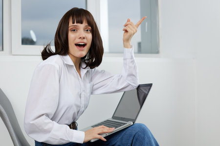 A young woman sits comfortably with a laptop, smiling broadly and pointing upward, capturing a moment of excitement and discovery in a bright home setting.の写真素材