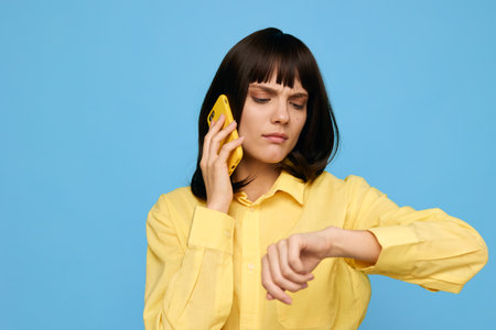 A young woman in a bright yellow shirt speaks on a mobile phone while checking her watch against a blue studio background, creating a dynamic everyday moment and a sense of urgency.の写真素材