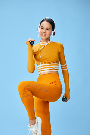 Young woman fitness athlete in bright orange workout set performs knee raise with dumbbells and headphones against a clean blue studio background, active sport pose.の写真素材