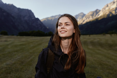 A cheerful young woman stands outdoors in a scenic mountain landscape. She wears a dark jacket and smiles softly, exuding calm confidence and curiosity as the setting enhances the moment.の写真素材