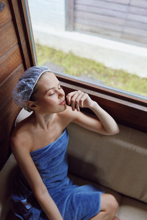 Relaxed woman wrapped in blue towel with hair cap enjoying calm moment near window, serene mood, natural light, wellness and spa concept, indoor setting.の写真素材