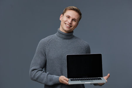 Smiling man wearing gray turtleneck sweater holding open laptop computer isolated on gray background...
