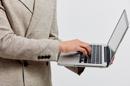 Professional young woman in beige blazer using silver laptop computer, standing against plain white...