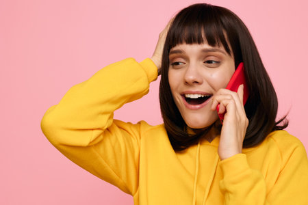 A cheerful young woman wearing a yellow hoodie talks on a red smartphone against a soft pink...