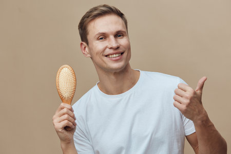 Smiling man with short brown hair holding a wooden hairbrush, showing thumbs up gesture, casual white t-shirt, isolated on beige background, studio portrait, positive emotion, lifestyle conceptの写真素材
