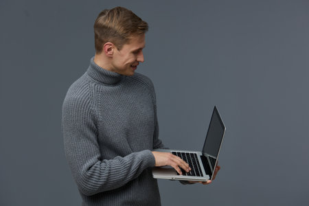 young smiling caucasian man wearing grey turtleneck sweater hold laptop look screen typing on keyboard isolated on dark grey background digital nomad conceptの写真素材
