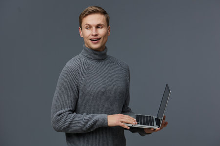 Smiling man holding laptop computer in hands, looking sideways, wearing cozy gray turtleneck, studio portrait on plain gray background, technology, work, casual style, indoor. People concept.の写真素材
