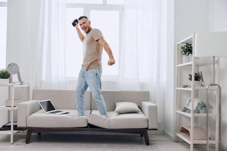 Energetic man celebrating success while standing on a couch, dressed casually in light clothing, with a joyful expression that reflects a modern lifestyle.の写真素材