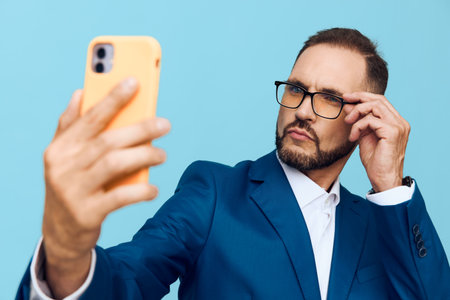A business professional man poses for a selfie against an isolated blue background, exuding confidence and focus while adjusting his glasses and checking his phone.の写真素材