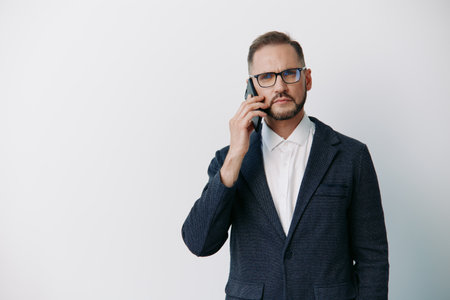 A business professional man speaks on a phone, standing against an isolated colored background, conveying confident focus and thoughtful emotion in a sleek corporate portrait.の写真素材