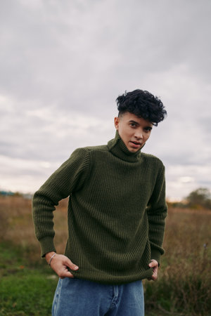 A young man stands in an open field wearing a chunky green turtleneck, relaxed pose, authentic everyday style, candid portrait under soft overcast light.の写真素材