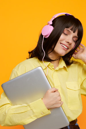A cheerful young woman wearing a pastel yellow shirt and pink headphones rests against a bright orange backdrop, holding a silver laptop and enjoying a relaxed music moment.の写真素材