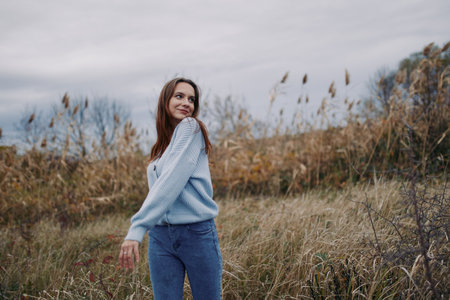 A relaxed portrait of a young woman in a cozy sweater walking through a grassy autumn field, radiating authenticity and natural confidence.の写真素材
