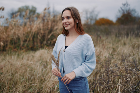 Young woman in a natural field wearing a cozy sweater, smiling and holding dried grasses, captured with authentic and credible lifestyle portraiture and warm candid expression.の写真素材