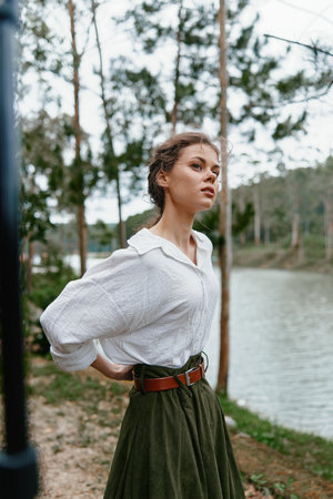 Young woman in a white blouse and green skirt standing by a river, exuding confidence and tranquility amidst a natural settingの写真素材