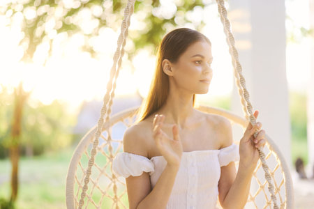 Young woman in white dress sitting on a swing, experiencing a moment of calm in a sunlit garden, showcasing peace and tranquility with natureの写真素材