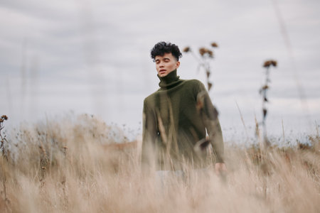 A young man in a knitted turtleneck walks through tall dry grass, a candid portrait emphasizing natural authenticity and credible, unposed presence beneath a muted, overcast sky.の写真素材