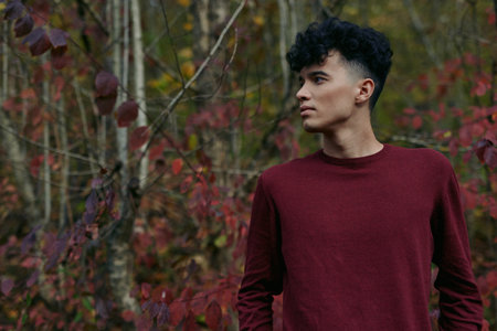 Young man in a burgundy sweater stands among autumn foliage, captured with natural light and authentic framing to convey genuine presence, subtle emotion and believable portrait realism.の写真素材