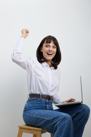 A joyful young woman sits on a wooden stool with a laptop in her lap, raising her fist in triumph, exuding motivation, success, and celebration in a bright studio setting.の写真素材