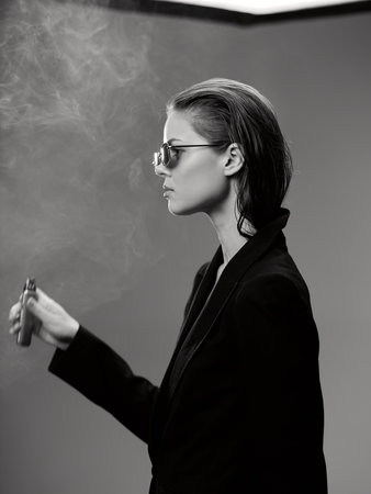 Black and white portrait of a stylish woman wearing sunglasses smoking with smoke around her.の写真素材