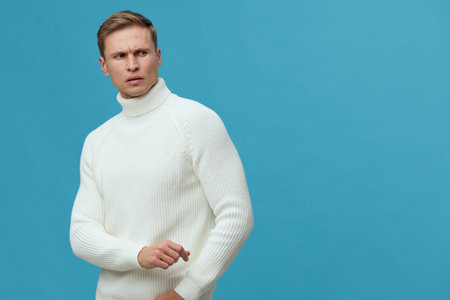 Thoughtful young man in white sweater looking sideways against plain blue background studio portrait, casual fashion, pensive mood, modern style, indoor setting. People lifestyle conceptの写真素材