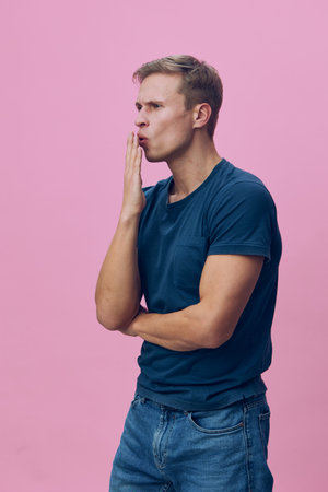 Thoughtful young man with short hair wearing casual blue t-shirt and jeans, isolated on pink...
