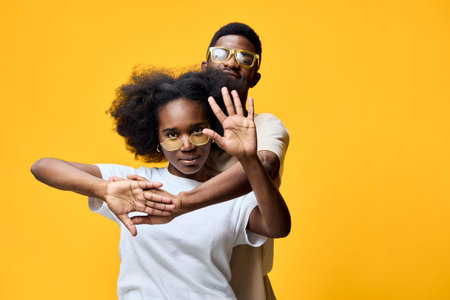 Young African man and woman posing together against a bright yellow background, showcasing playful emotions and modern fashion styleの写真素材