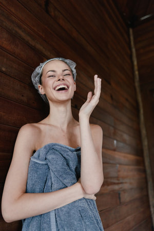 laughing woman wrapped in towel with hair covered standing against wooden wall enjoys spa moment and relaxation, natural skin, wellness conceptの写真素材