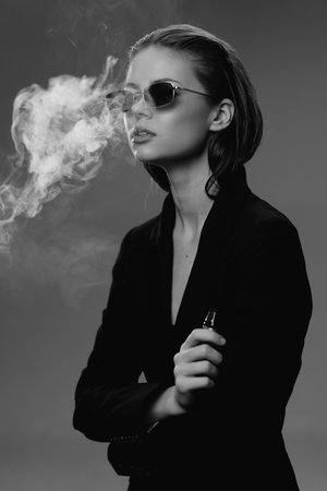 Black and white portrait of a woman vaping with sunglasses in a dark outfit, exhaling smoke in a...