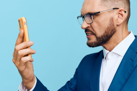 A professional man in a suit checks his phone with focused emotion against a solid blue background, conveying confidence, concentration, and modern business readiness.の写真素材