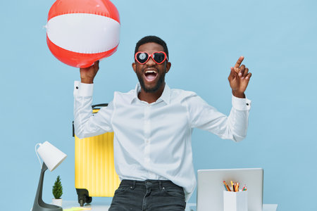 Joyful man celebrating summer vacation holding striped beach ball, wearing sunglasses and casual white shirt in modern colorful office with travel accessories. Happiness, leisure, relaxation concept.の写真素材