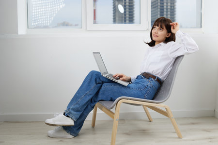 A relaxed female professional sits in a bright, modern room with a laptop, creating a calm and productive mood. She wears denim jeans and a white shirt, leaning back in a simple chair.の写真素材