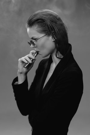 Black and white portrait of a thoughtful woman in sunglasses vaping with a dark background and...