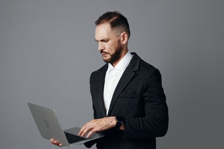 A focused professional man in a dark suit uses a laptop against an isolated gray background, conveying confidence, concentration, and business readiness.の写真素材
