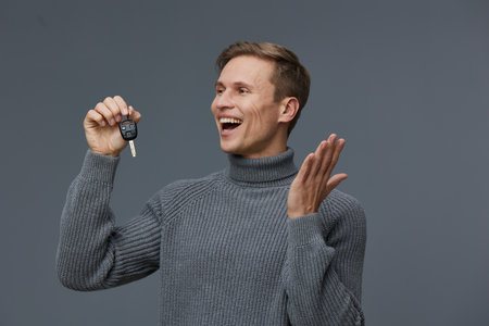 Smiling man holding car key with happy expression, wearing casual gray sweater, against plain gray background. Portrait of a joyful person celebrating new vehicle purchase or achievement, lifestyle concept.の写真素材