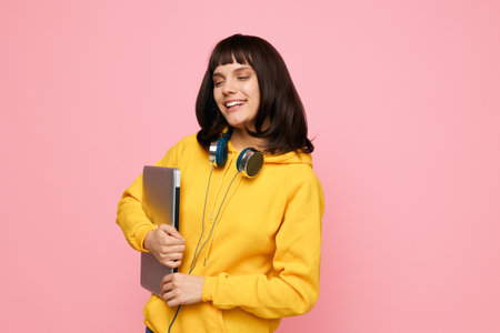 A cheerful young woman wearing a bright yellow hoodie holds a laptop with headphones resting around her neck, standing against a soft pink backdrop, exuding energy and creative mood.の写真素材