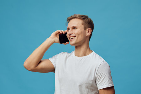 smiling caucasian man talking on smartphone using voice assistant technology wearing white t-shirt isolated on blue background casual lifestyle conceptの写真素材