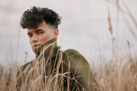 Moody outdoor portrait of a young man in a chunky green sweater among tall dry grass, capturing a candid natural expression that emphasizes authenticity and visual credibility in a realistic scene.の写真素材
